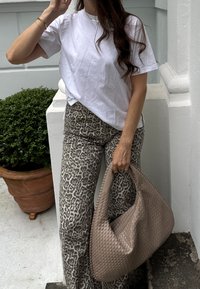 Bílý krátký rukáv tričko v kombinaci se světlými šedými kalhotami s leopardím vzorem a vysokým pasem; drží tkanou béžovou kabelku typu hobo.