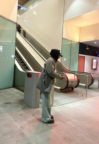 Scale grigia con lati metallici, affiancata a una persona che indossa una giacca grigia e pantaloni larghi chiari, vicino a un cestino della spazzatura argentato.