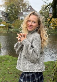 Grå melert strikkegenser med høy hals, løse ermer og cropped snitt, kombinert med et rutete skjørt, i en naturlig utendørs setting.