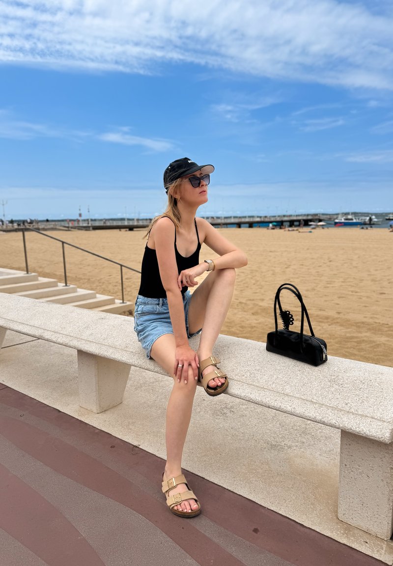 Giovane donna con occhiali da sole, canotta nera e pantaloncini di jeans seduta su una panchina vicino alla spiaggia con una borsa nera nelle vicinanze.