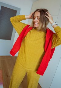 Femme souriante, les mains dans les cheveux, portant un pull moutarde et un sweat-shirt rouge posé sur les épaules, debout dans une pièce lumineuse.