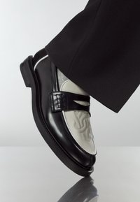 Black leather loafers with a textured grey leather panel, featuring a strap detail and a low, flat sole. Smooth and polished finish.