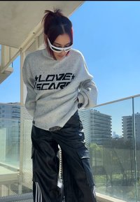Suéter gris de punto con el texto "LOVE SCAR" en negro, combinado con pantalones cargo negros brillantes con rayas blancas y el logo de Adidas, de corte relajado.