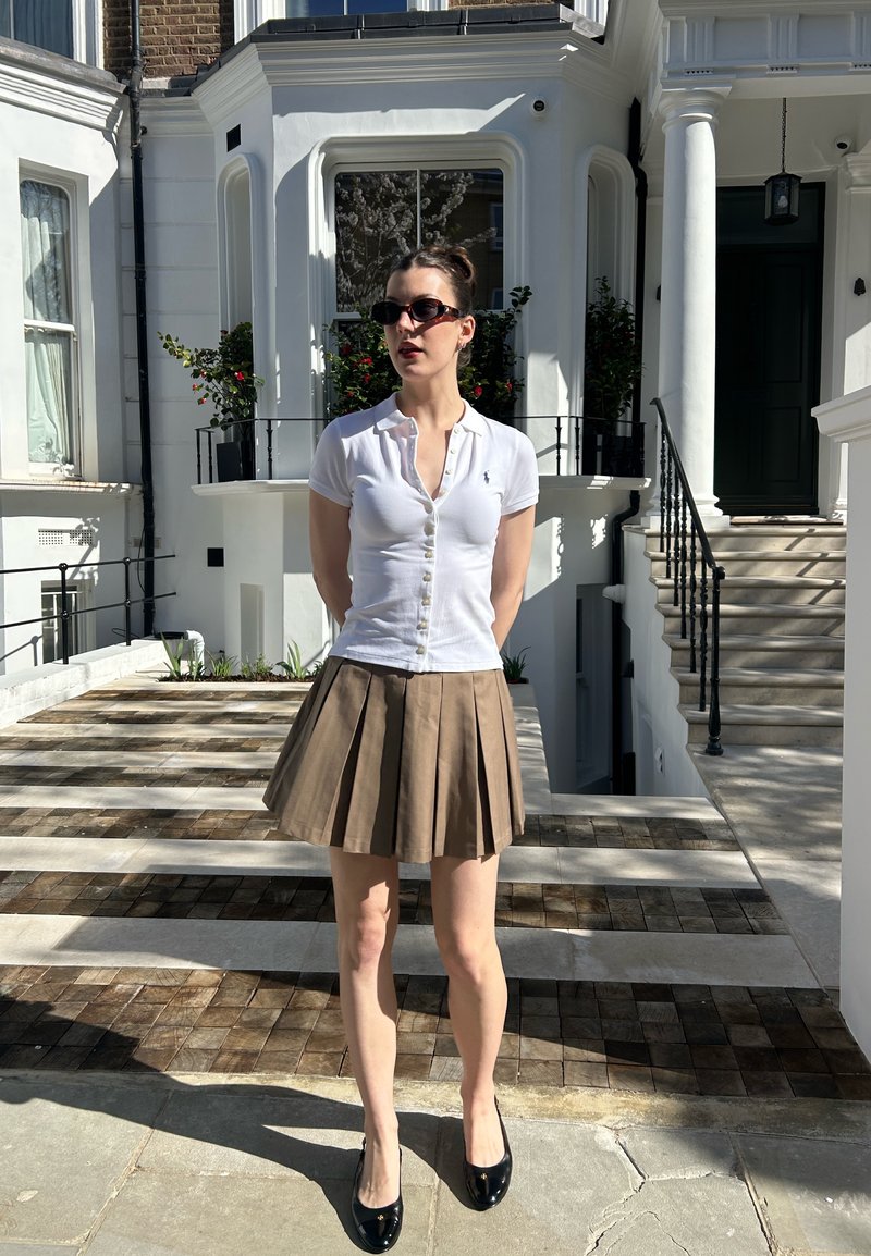 Donna con occhiali da sole, camicia bianca con bottoni, gonna plissettata marrone e scarpe nere in piedi davanti a un edificio bianco con scale e piante.