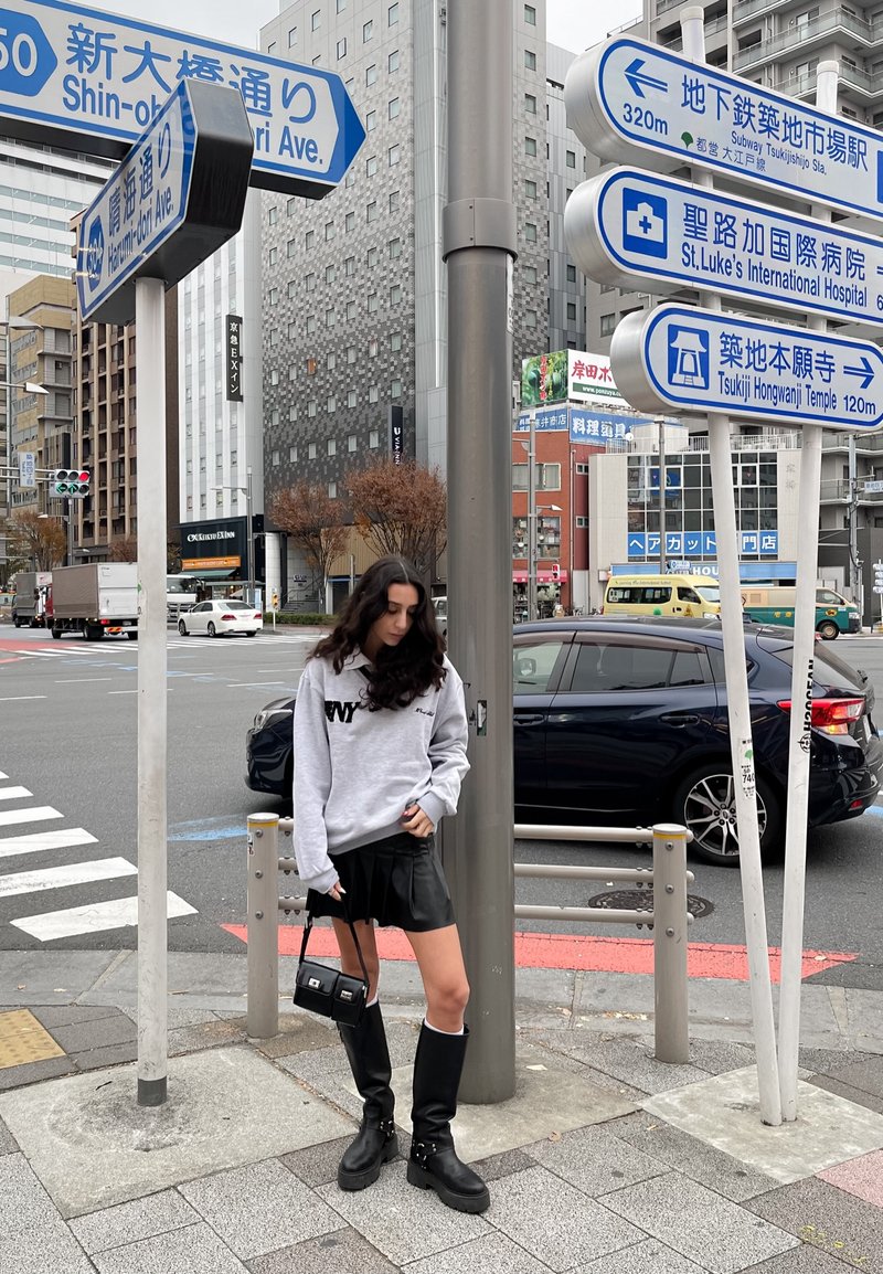 Γκρι φούτερ με εκτύπωση "ΝΥ", μαύρη δερμάτινη μίνι φούστα, μαύρες μπότες μέχρι το γόνατο με αγκράφες, στέκεται δίπλα σε πινακίδες δρόμου σε αστική περιοχή.