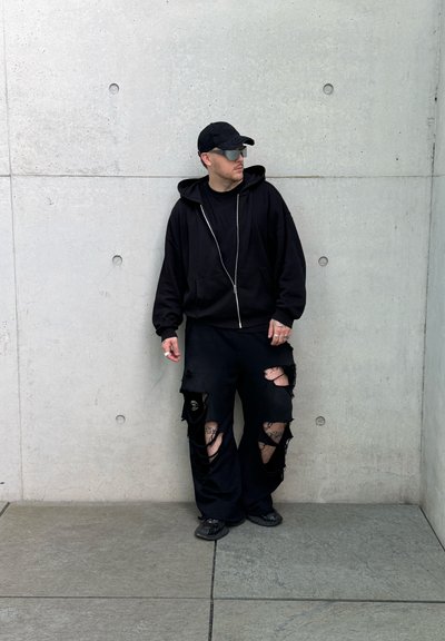 Hombre con gorra negra, gafas de sol, sudadera negra con capucha y pantalones negros rotos con tatuajes visibles, de pie contra una pared de hormigón gris.
