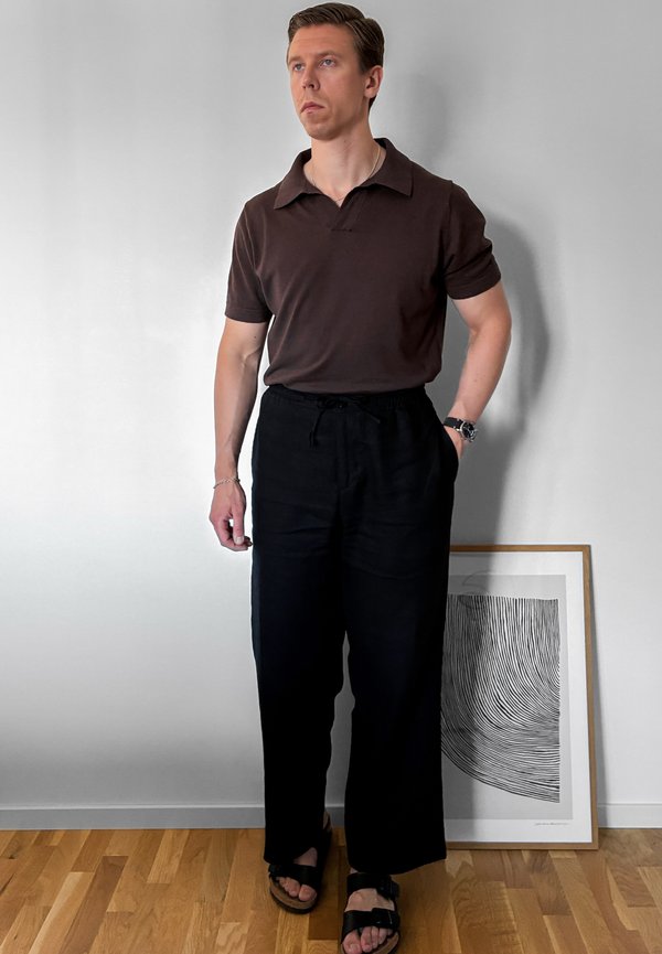 Man wearing a brown polo shirt, black wide pants, and black sandals standing by a framed black and white abstract artwork on wooden floor.