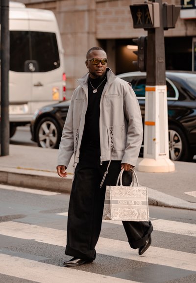 Chaqueta gris con cremallera, cuello de tortuga negro, pantalones anchos negros, zapatos negros elegantes, gafas de sol amarillas, sosteniendo un bolso con estampado de Christian Dior.