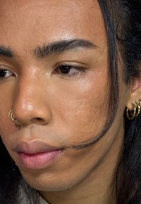 Close-up van een gezicht met een gladde, lichte huid, bruine ogen, subtiele lipkleur en een gouden oorring. Heeft fijne haardtextuur en een lichte glans.