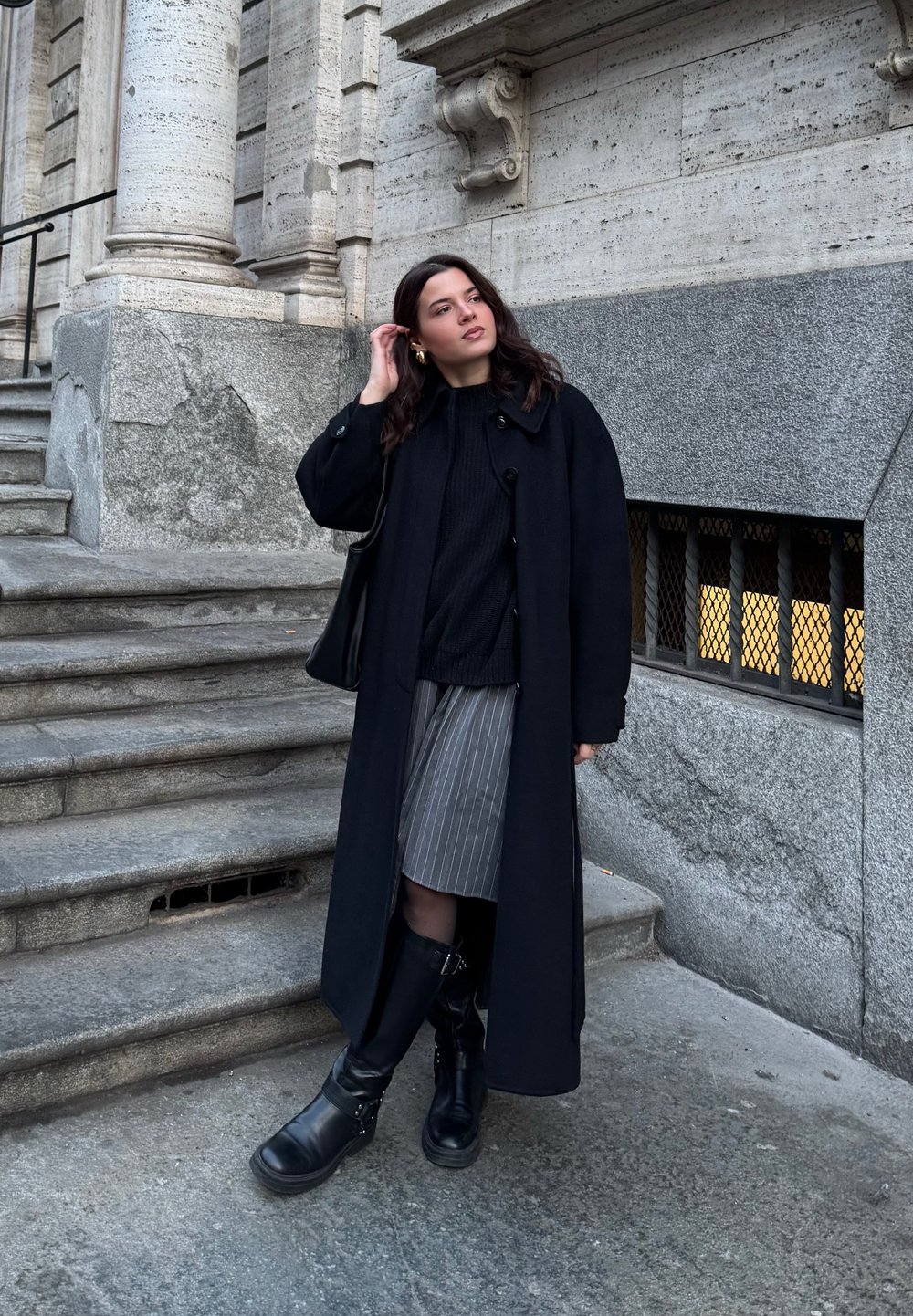Lungo cappotto nero su un maglione nero a trama, abbinato a una gonna a righe e stivali neri fino al ginocchio. In piedi su gradini di pietra.