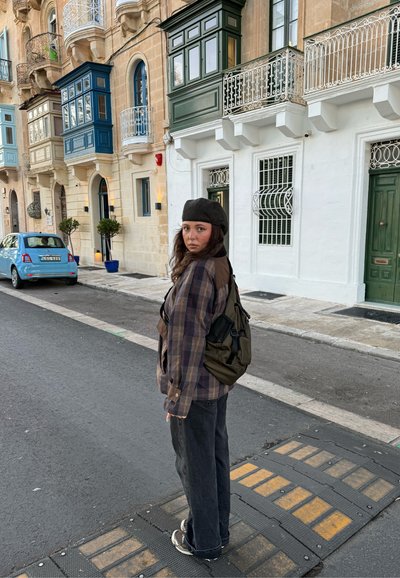 Joven mujer con camisa de cuadros, boina negra y mochila, de pie en la calle con balcones tradicionales coloridos y un coche azul estacionado cerca.