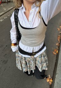 Szürke trikó fehér-kék csíkos gombos ing felett, kockás szoknyával párosítva. Fekete öv és kiegészítők fokozzák a rétegezett megjelenést.