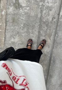 Mustad musta võrkmaterjaliga kingad laiate rihmadega, kantud musta seeliku ja valge T-särgiga, millel on punane graafiline tekst, paigutatud hallile betoonpinnale.