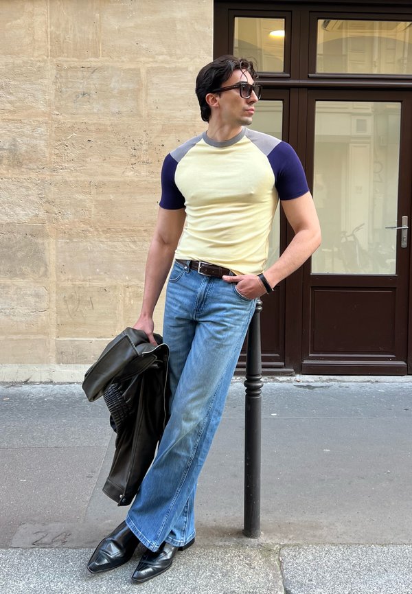 Uomo con occhiali da sole, t-shirt gialla e blu navy, jeans blu e stivali neri si appoggia a un palo mentre tiene una giacca di pelle nera all'aperto.