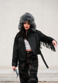 Černá kožená bunda s třásněmi, nošená přes bílý crop top. Šed�á kožešinová kapuce, černé kalhoty se vzorem a sluneční brýle v ruce.