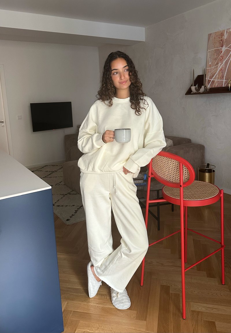 Ensemble en sweat-shirt et pantalon de survêtement de couleur crème avec un tissu texturé. Tasse élégante à la main, se tenant à côté d'une chaise rouge avec un siège en osier.