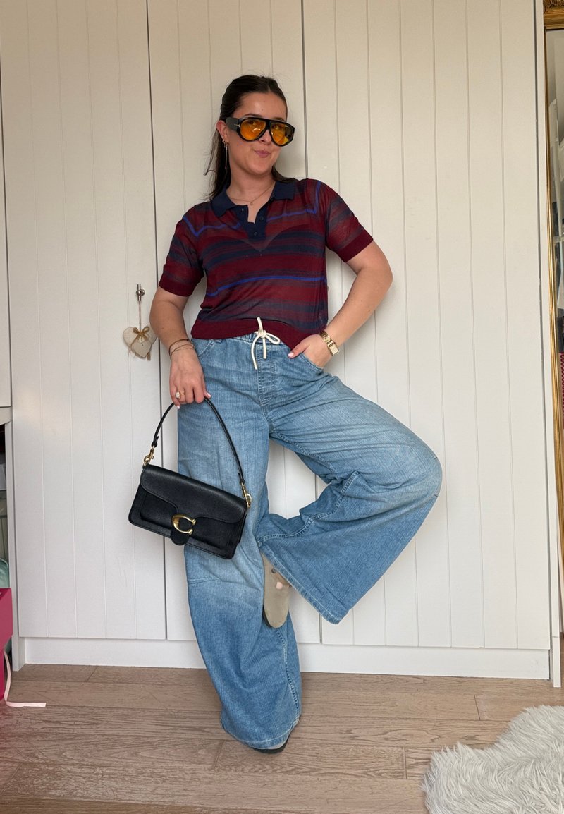 Woman posing against white paneled wall wearing striped maroon and blue polo shirt, wide-leg jeans, orange-tinted sunglasses, and holding black handbag.