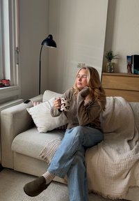 Przytulna scena w salonie przedstawia kobietę siedzącą na jasnoszarym sofie, trzymającą białą filiżankę w czerwone kropki, ubraną w brązowy sweter i niebieskie dżinsy.