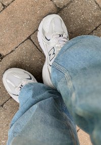 Zapatillas blancas de New Balance con acentos de malla y cuero, exhibidas junto a unos pantalones de mezclilla azul claro sobre un pavimento de piedra.