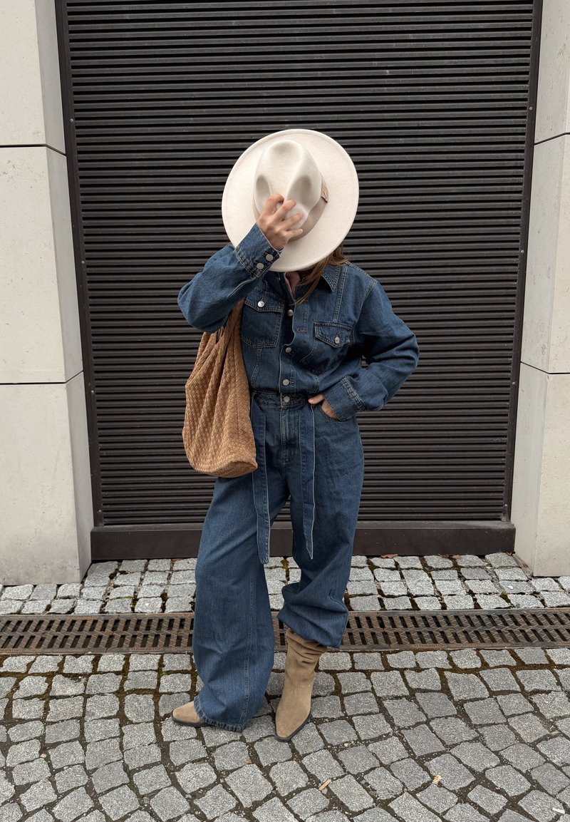 Personne en veste en jean et pantalon avec des bottes couleur sable tient un chapeau blanc à large bord couvrant le visage, portant un sac à bandoulière marron texturé dans une rue pavée.