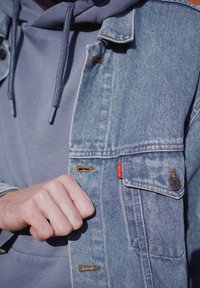 Vaaleansininen farkkutakki, jossa on punainen logotunniste, sivutaskut ja ruskea ompelu. Päälle puettu harmaa huppari, jossa on kiristysnauhat.