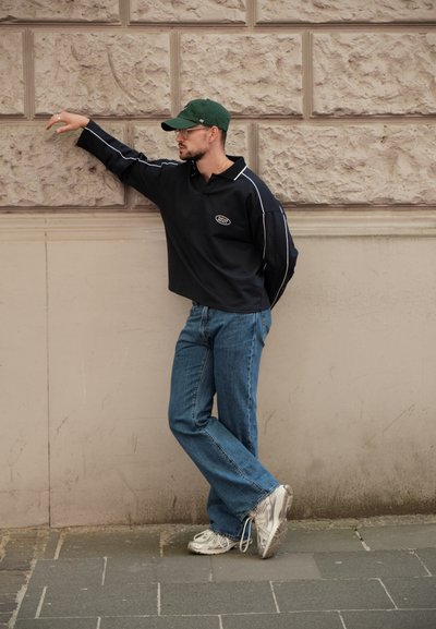 Polo negro de manga larga con acentos blancos, jeans acampanados azules y zapatillas blancas. Persona apoyada contra una pared texturizada en un entorno urbano.