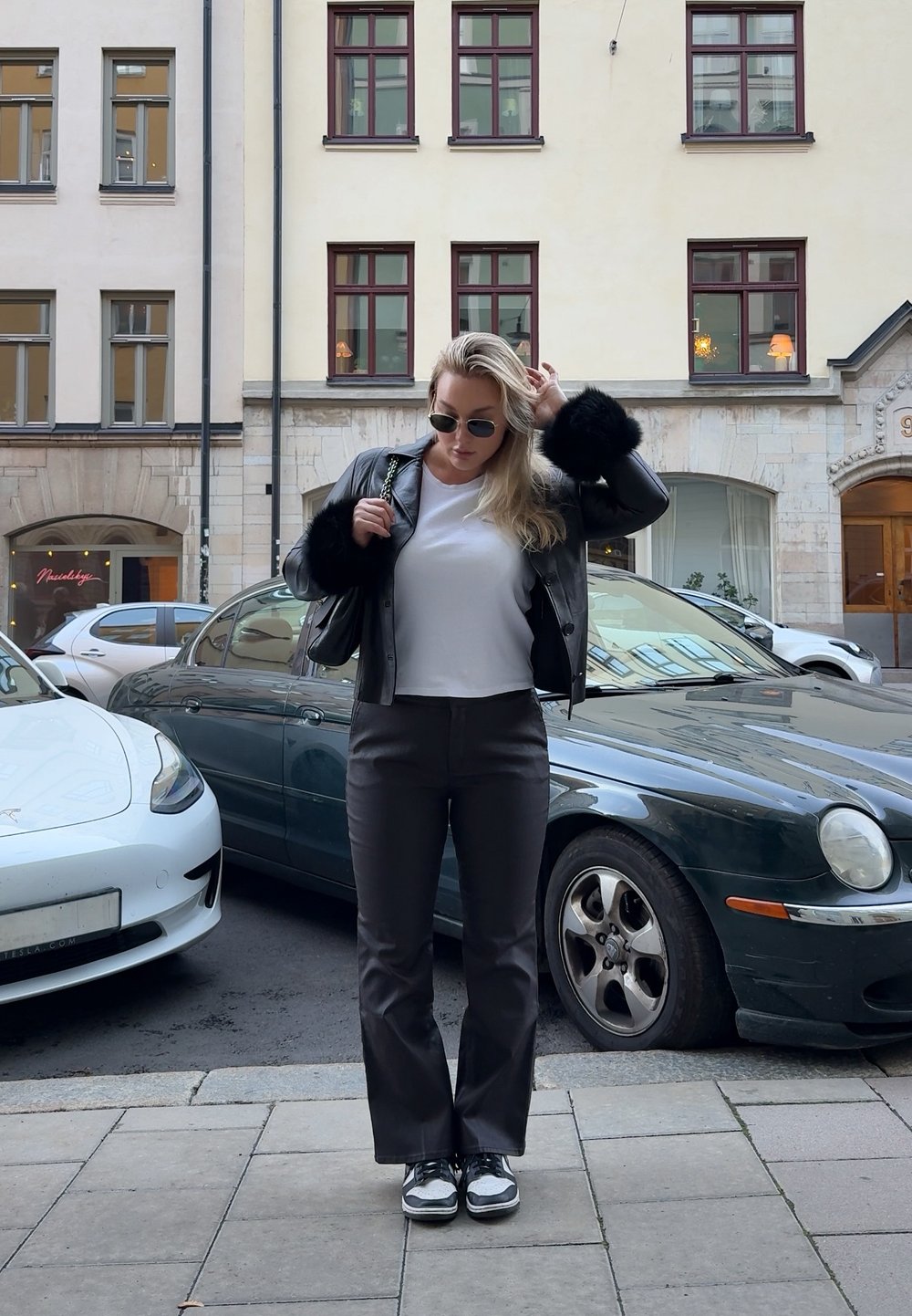 Czarna skórzana kurtka z futrzanymi akcentami, biała koszulka, szare spodnie i czarno-białe sneakersy. Miejska sceneria z zaparkowanymi samochodami.