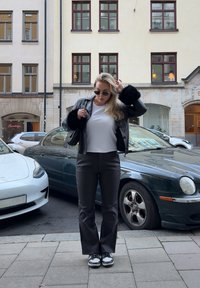 Czarna skórzana kurtka z futrzanymi akcentami, biała koszulka, szare spodnie i czarno-białe sneakersy. Miejska sceneria z zaparkowanymi samochodami.