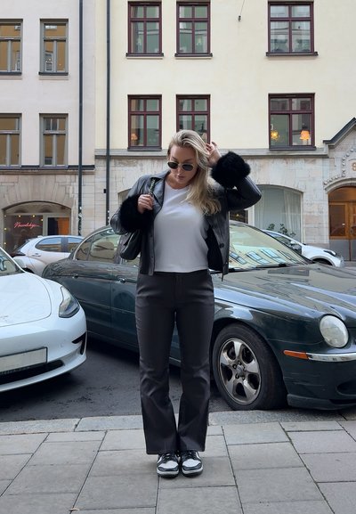 Chaqueta de cuero negra con detalles de piel, camiseta blanca, pantalones grises y zapatillas negras y blancas. Entorno urbano con coches aparcados.