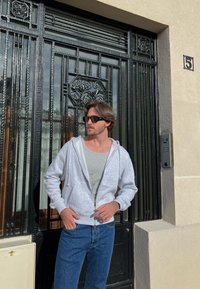 Szürke zipzáros kapucnis pulóver egy világosszürke póló felett, kék farmerrel párosítva. Fekete napszemüveget visel, és a háttérben egy textúrált ajtó látható.