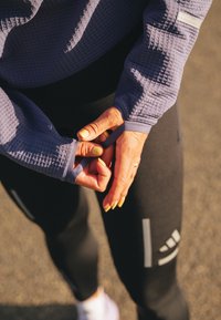 Paarse, gestructureerde lange mouw top met duimgaten; strakke zwarte legging met een reflecterend logo. Zichtbare licht- en schaduweffecten.