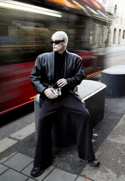Chaqueta de cuero negra sobre un cuello de tortuga negro, pantalones anchos negros, bolso plateado y gafas de sol. Borroso por el movimiento de un vehículo que pasa.