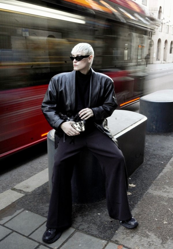 Black leather jacket over a black turtleneck, wide-legged black trousers, silver bag, and sunglasses. Motion blur from a passing vehicle.