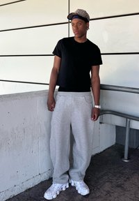 Black short-sleeve t-shirt, grey oversized sweatpants with pockets, and white chunky sneakers; wearing a patterned cap and wristwatch.