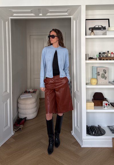 Mujer con pelo rizado que lleva gafas de sol, cárdigan azul claro, top negro, pantalones cortos de cuero marrón y botas negras hasta la rodilla en una habitación moderna.