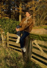 Brun læderjakke, ternet tørklæde, blå jeans og hvide sneakers. Sidder på et slidt træhegn midt i efterårsblade.