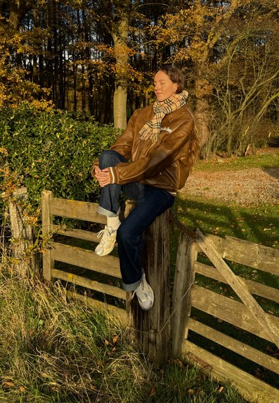 Chaqueta de cuero marrón, bufanda de cuadros, jeans azules y zapatillas blancas. Sentado en una cerca de madera envejecida entre el follaje otoñal.
