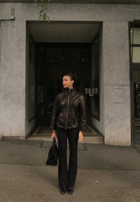 Giacca di pelle marrone con colletto alto, pantaloni neri a zampa d'elefante e borsa nera. In piedi davanti a un edificio con pareti in pietra grigia.