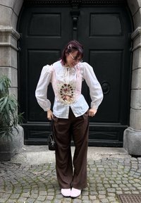Blusa bianca con maniche a sbuffo, corsetto rosa con grafica di un gatto, pantaloni ampi marroni e scarpe rosa, completati da una piccola borsa nera.