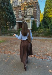 Bruine maxi rok met plooien, lichtblauwe blouse en een bruine handtas met een patroon. Rode bakstenen gebouw en groene bladeren op de achtergrond.