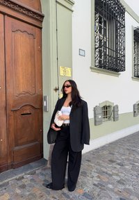 Oversized donker colbert, wit tanktopje, zwarte wijde broek, houdend een crèmekleurige clutch. Steenstraten en houten deur op de achtergrond.