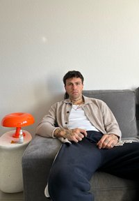 Μπεζ ζακέτα πάνω από λευκό πουκάμισο, ναυτικές φόρμες, τατουάζ ορατά στα χέρια. Πορτοκαλί λάμπα επιτραπέζια δίπλα σε ένα πλευρικό τραπέζι με υφή σε φωτεινό χρώμα.