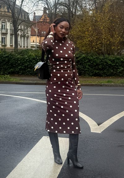 Vestido midi marrón con lunares, de manga larga y silueta ajustada, combinado con botas negras de tacón hasta la rodilla y un bolso negro.