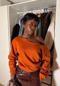 Femme aux cheveux courts portant un haut orange décolleté et une jupe en cuir marron se tient devant une armoire ouverte, tenant un sac à main.