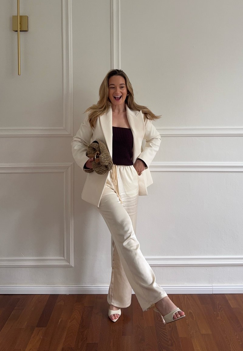 Femme souriante, portant un blazer crème et un pantalon avec un haut noir, tenant un sac tressé, debout sur un sol en bois devant un mur blanc à panneaux.