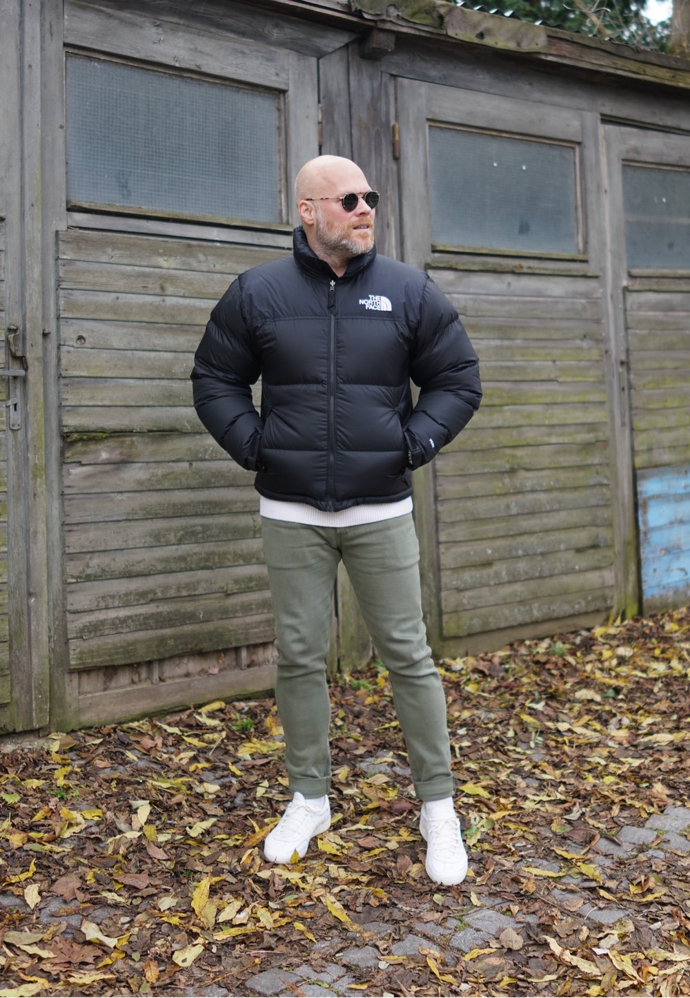Zwarte pufferjack met een hoge kraag, ritssluiting en logo. Gecombineerd met lichtgroene broek en witte sneakers. Achtergrond van houten structuren.