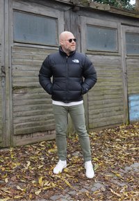 Zwarte pufferjack met een hoge kraag, ritssluiting en logo. Gecombineerd met lichtgroene broek en witte sneakers. Achtergrond van houten structuren.