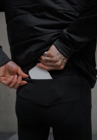 Leggings de sport noirs avec une poche zippée, présentant une coupe ajustée. Un objet blanc, probablement un téléphone, est en train d'être glissé dans la poche.