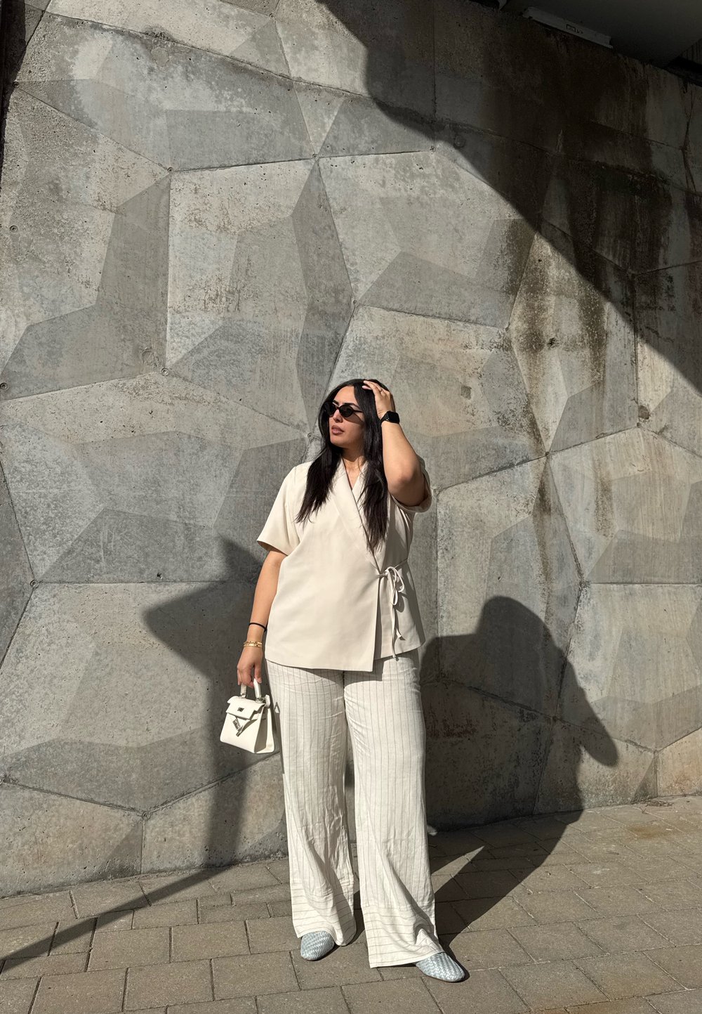 Beige wikkeltop met korte mouwen, witte gestreepte wijde broek en een kleine witte handtas. Achtergrond heeft een textuur van een betonnen muur.