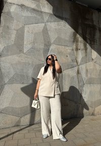 Beige wikkeltop met korte mouwen, witte gestreepte wijde broek en een kleine witte handtas. Achtergrond heeft een textuur van een betonnen muur.